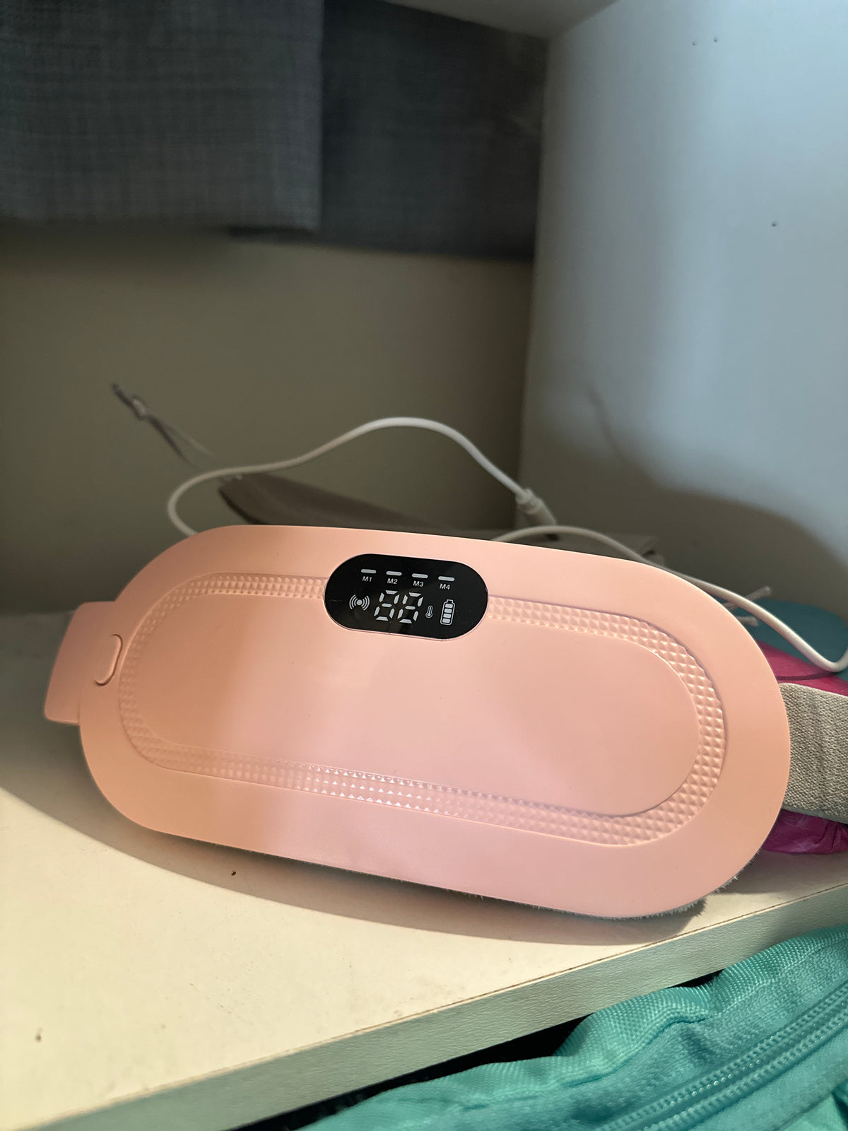 A sleek pink electronic device featuring a digital display, placed on a clean white surface beside a soft gray drawstring pouch and subtle background accents.