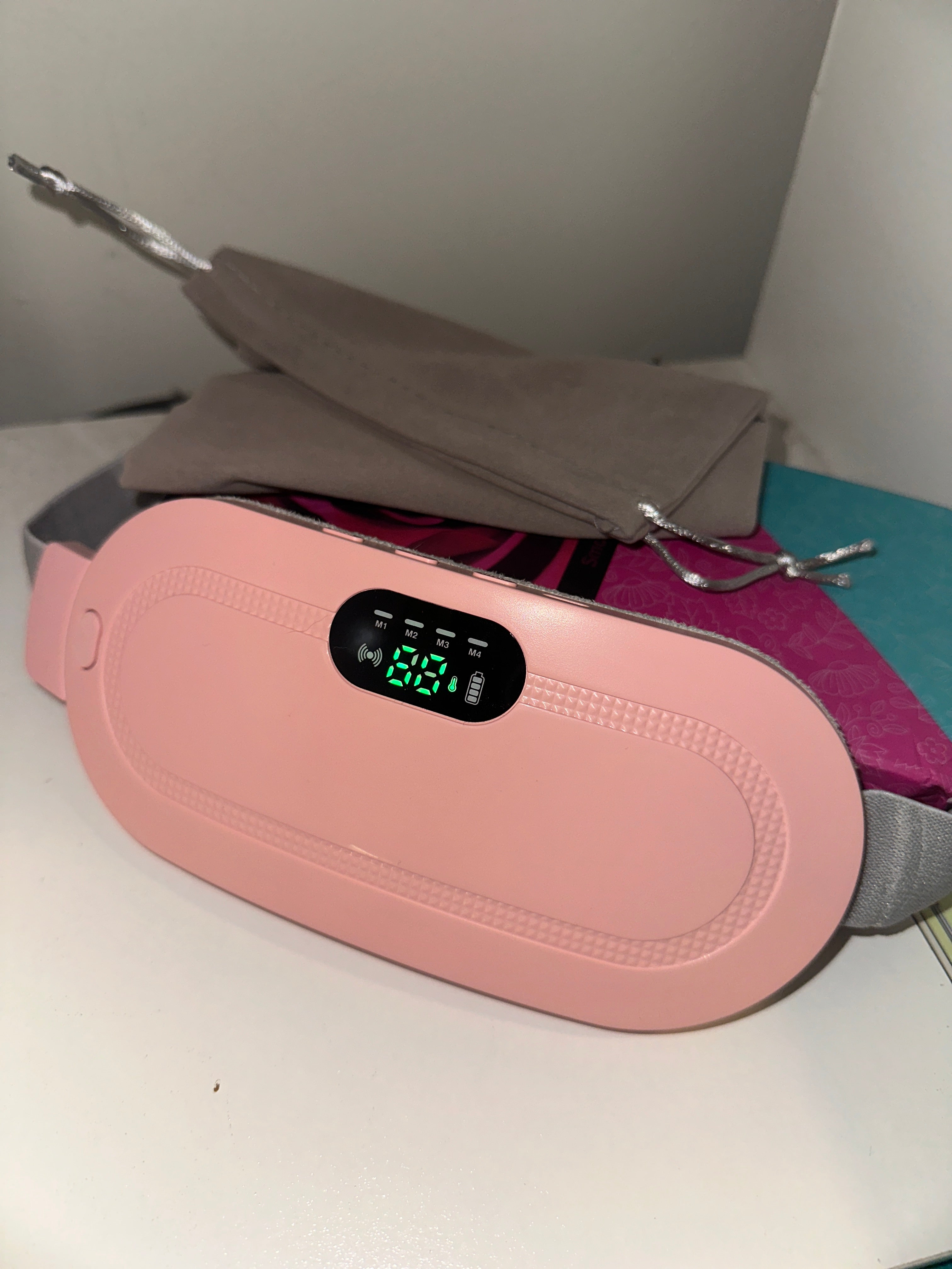 A sleek pink electronic device featuring a digital display, placed on a clean white surface beside a soft gray drawstring pouch and subtle background accents.