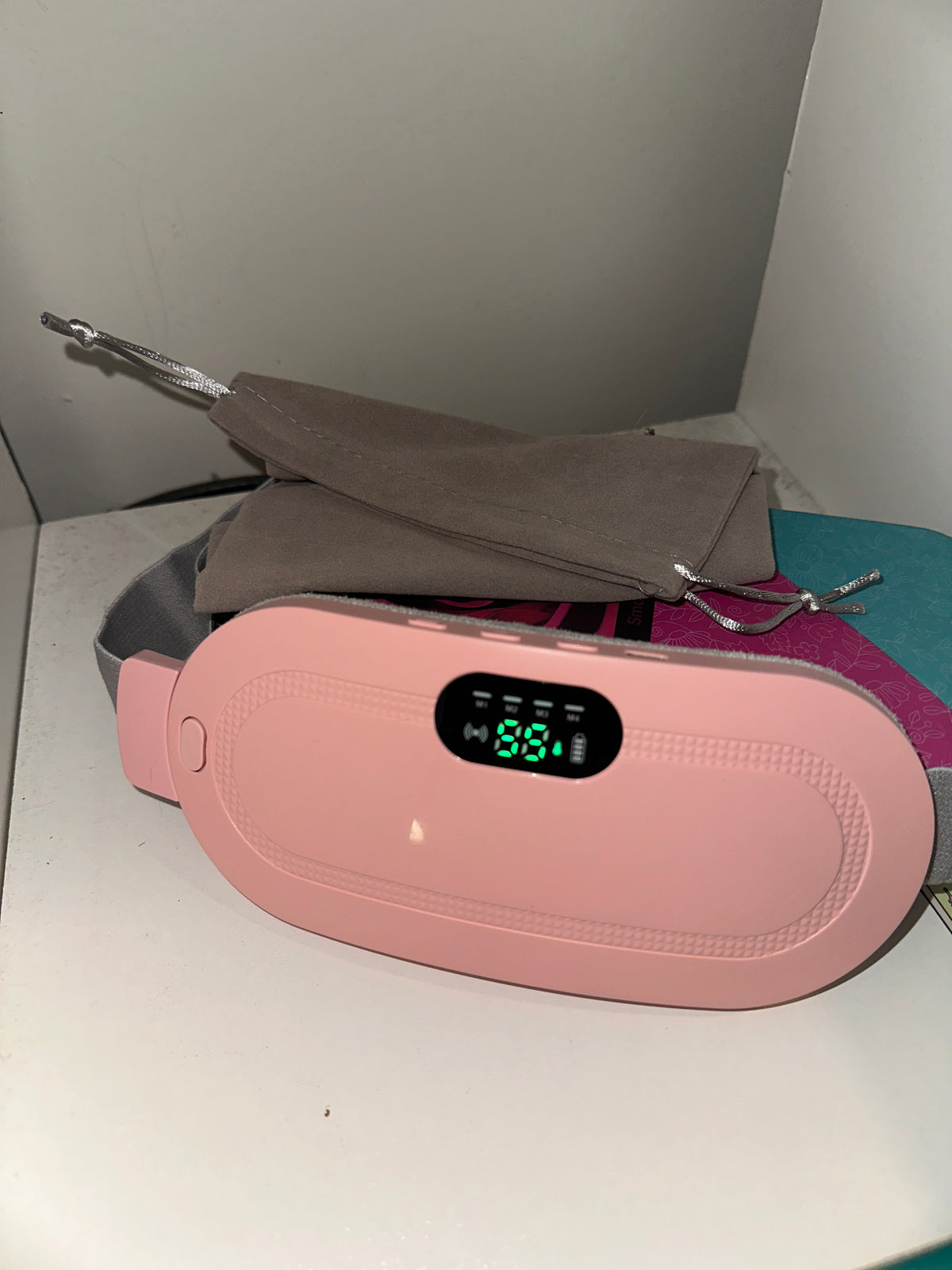 A sleek pink electronic device featuring a digital display, placed on a clean white surface beside a soft gray drawstring pouch and subtle background accents.