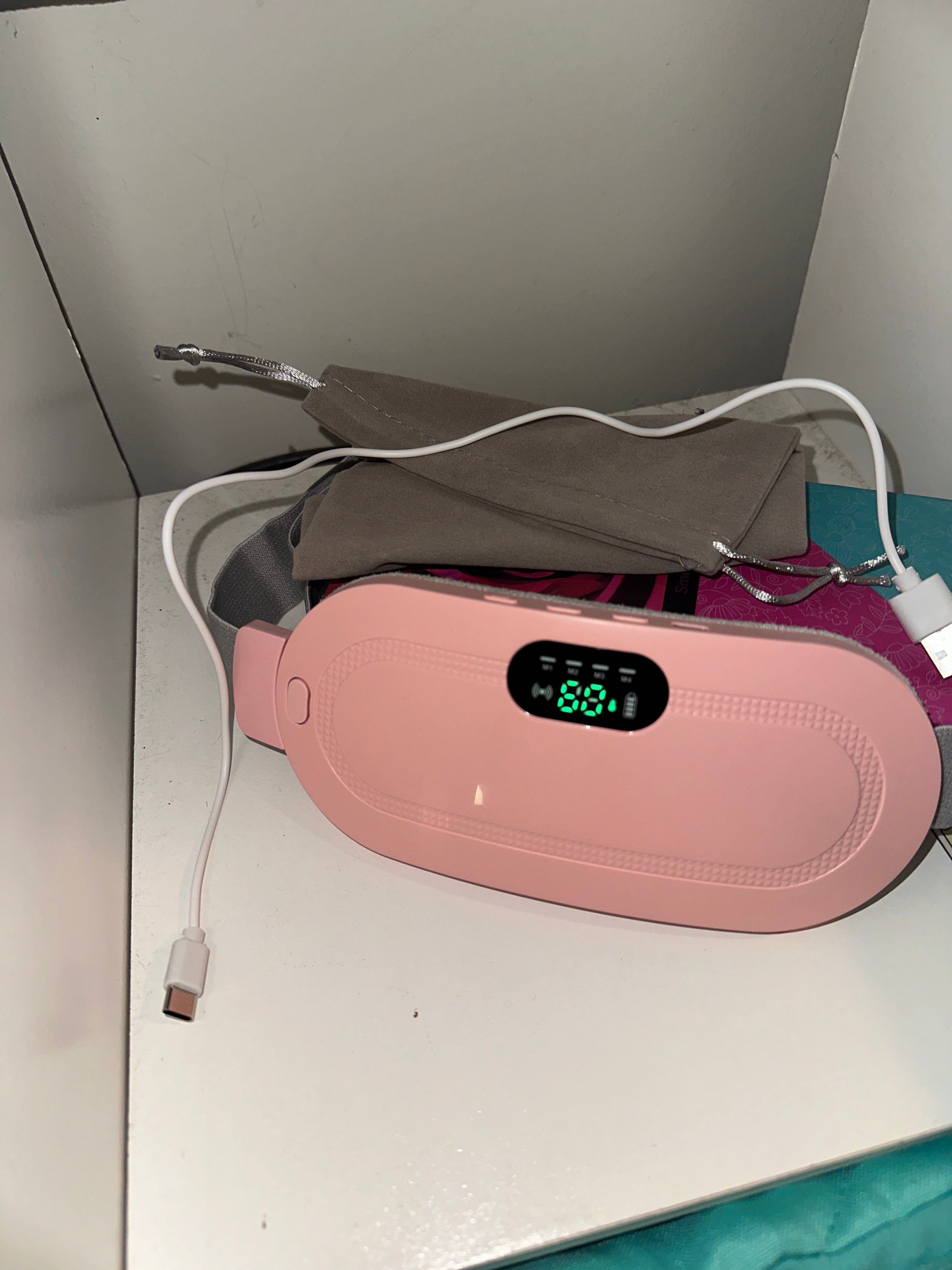A sleek pink electronic device featuring a digital display, placed on a clean white surface beside a soft gray drawstring pouch and subtle background accents.